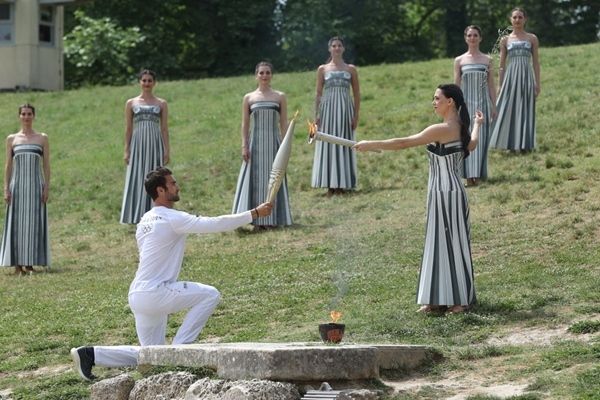 扮演最高女祭司的演員瑪麗·米娜（前右）點燃第一棒火炬手——希臘運動員、東京奧運會男子賽艇項目金牌得主斯特凡諾斯·恩圖斯科斯手中的火炬。新華社記者 趙丁喆 攝